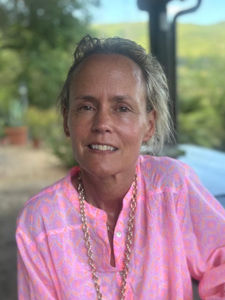 Lulu Townsend smiling on a sunlit terrace in a pink blouse, green hills soft in the distance.